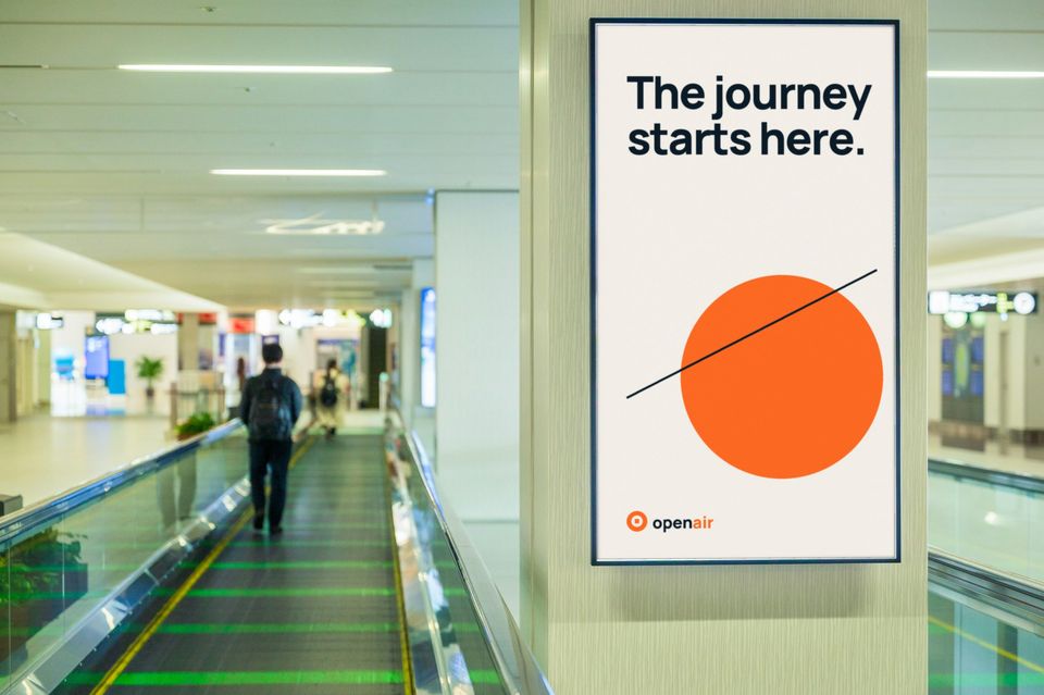 A traveller on an airport walkway passes a portrait screen showing an orange sun and the line "The journey starts here."