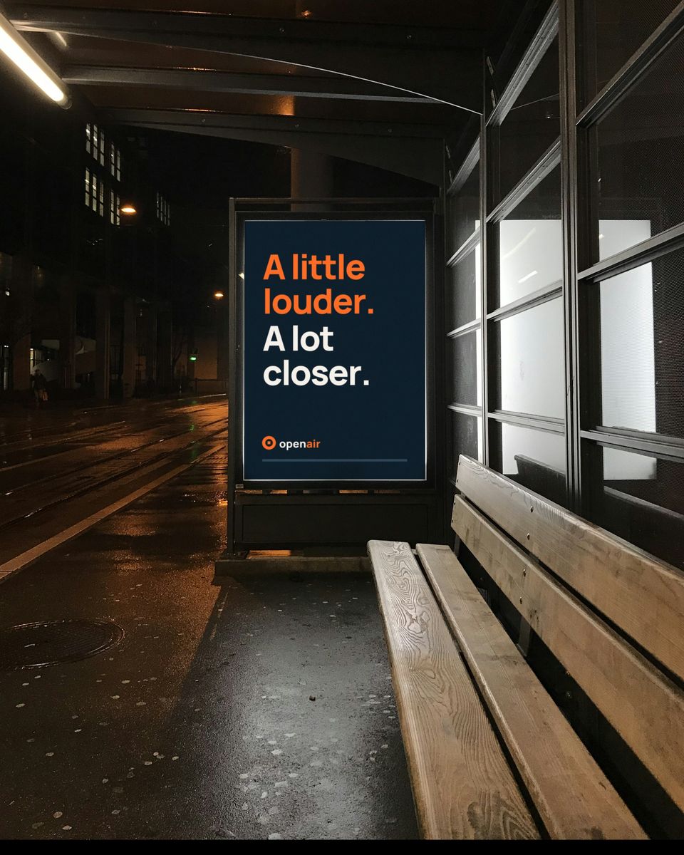 A lit bus-shelter panel at night on a wet street, showing a navy poster that reads "A little louder. A lot closer."