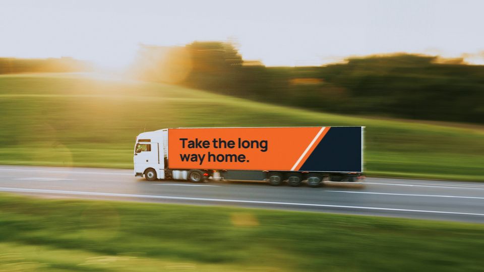 A lorry crossing open countryside at sunset with an orange trailer wrap reading "Take the long way home."