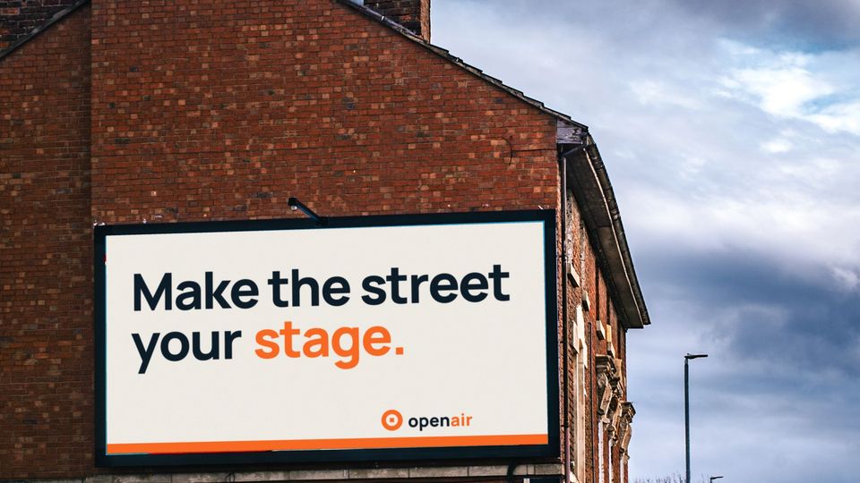 A large roadside billboard on the gable end of a red-brick building, showing an Open Air Media House concept poster