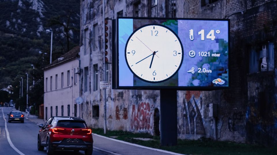 A roadside digital screen at dusk showing the time and the weather above a quiet street