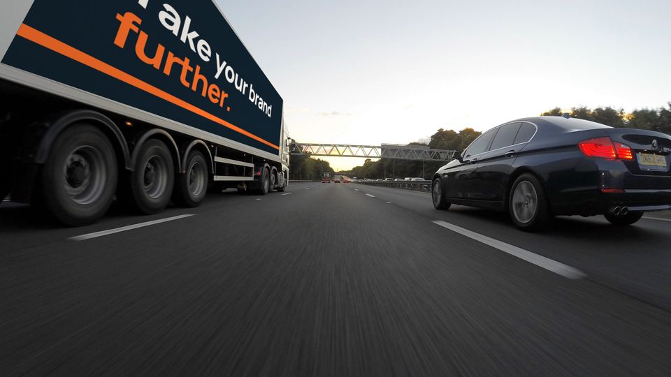 A lorry on a UK motorway at dusk with a navy and orange trailer wrap reading "Take your brand further."